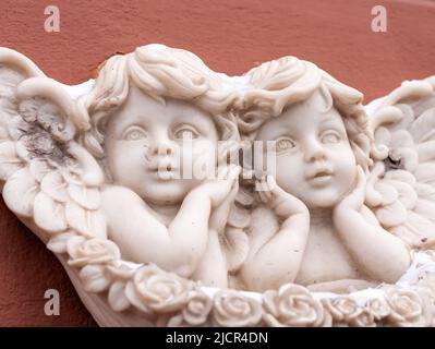 Zwei kleine Engel mit Flügeln Marmorskulptur an der roten Wand des Gebäudes. Nahaufnahme, Vorderansicht. Stockfoto