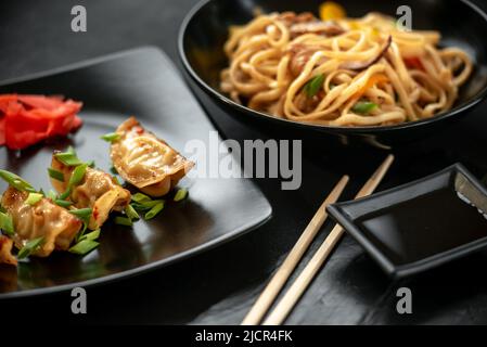 Set asiatischer Speisen auf dunklem Hintergrund auf schwarzem Teller. Japanische frittierte Gyoza-Knödel und udon-Nudeln Stockfoto
