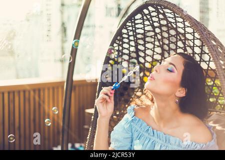 Eine junge Frau mit hellem Make-up sitzt in einem Sommer-Café im Freien in einem Hängesessel und bläst Seifenblasen. Stockfoto