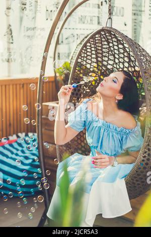 Eine junge Frau mit hellem Make-up sitzt in einem Sommer-Café im Freien in einem Hängesessel und bläst Seifenblasen. Stockfoto