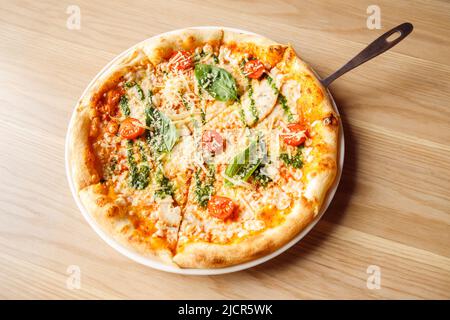 Italienische Pizza mit Huhn, Kirschtomaten, Mozzarella und Parmesan, Basilikum auf dem Teller mit Servierspatel auf Holztisch. Draufsicht, Lifestyle Stockfoto