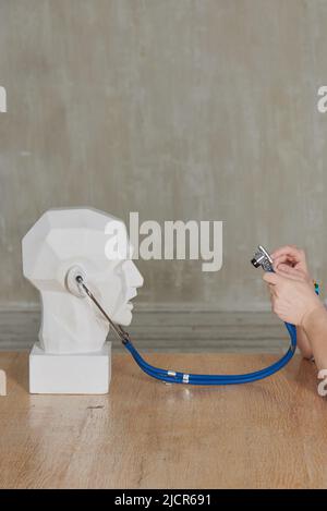 Der Kopf eines Gipsmannes mit einem Stethoskop in den Ohren und den Händen einer Frau an einer grauen Wand. Stockfoto