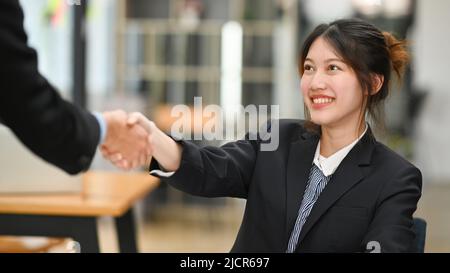 Lächelnde asiatische Geschäftsfrau schüttelt nach Abschluss eines Meetings oder eines Investitionsabschlusses ihre Hände mit ihrem Geschäftspartner Stockfoto