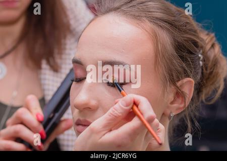 Eine Hand mit einem Pinsel malt das Augenlid eines Mädchens mit einer großen Handfläche. Weibliche Meisterin macht einer jungen Frau Make-up. Geschäftskonzept - Schönheitssalon, Gesichtshautpflege, Kosmetik. Stockfoto