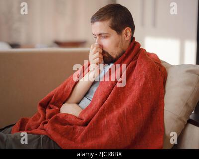 Coronavirus. Kranker Mann mit Corona-Virus, der sich auf dem Sofa niederlegt und sich von einer Krankheit zu Hause erholt. Seine Nase bläst. Das Konzept der hohen Temperaturen. Die Person ist schwer krank. Stockfoto
