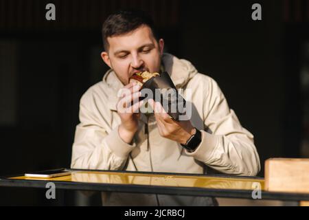 Schöner Mann, der während der Ruhepause traditionelle Hotdog-Küche serviert. Kaukasischer Mann, der draußen Straßenküche genießt. Männlich Essen ungesunde Fast Food Stockfoto