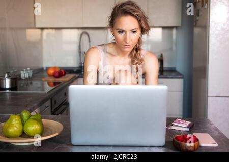 Junge kaukasische schöne schlanke blonde Frau sitzt tagsüber in der Küche an einem Tisch vor einem Laptop. Stockfoto