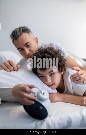 Ein süßer dunkelhaariger Junge, der während des Schlafes auf die Uhr schaut Stockfoto