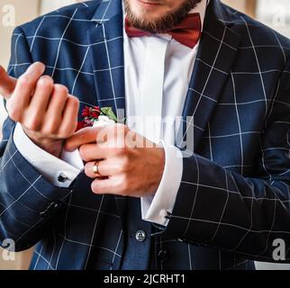 Verschwommener Hochzeitshintergrund. Ein junger Mann in einem karierten Anzug strafft seine Hemdmanschetten. Der Bräutigam in einer blauen Jacke strafft den Manschettenknauf am Ärmel Stockfoto