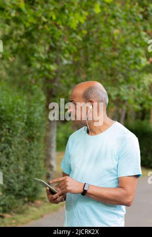 Seitenportrait eines haarlosen Mannes, der mit Kopfhörern auf seinem Handy in der Natur Musik hört, während er durch die Landschaft geht Stockfoto