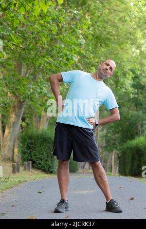 Ganzkörper-Unterbild eines erwachsenen Mannes, der sich dehnt und auf einem konkreten Weg zwischen der grünen Natur trainiert Stockfoto