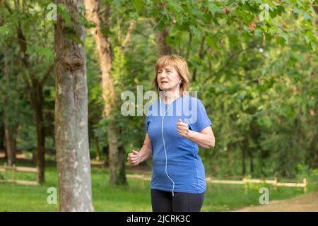 Ältere Dame, die in Sportkleidung durch die Natur geht, während sie mit Kopfhörern von ihrem Mobiltelefon Musik hört Stockfoto