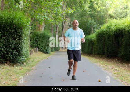 Frontportrait im Landschaftsformat eines älteren Mannes, der auf einem Pfad zwischen den grünen Bäumen eines Parks läuft Stockfoto