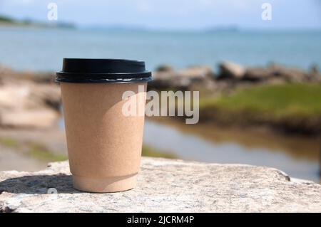 Papierkaffeetasse auf Felsen mit verschwommenem Strandhintergrund. Speicherplatz kopieren Stockfoto