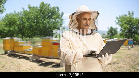 Reifer männlicher Bienenzüchter in Uniform mit einem Laptop-Computer auf einem Bienenstock Stockfoto