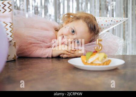 Niedliche kleine Geburtstag-Mädchen posiert in der Nähe Geburtstagskuchen in schönen rosa Festkleid. Horizontales Foto. Mädchen feiert Geburtstag Konzept Stockfoto