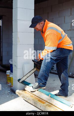 Vertikaler lächelnder Arbeiter in orangefarbener Schutzhülle mit Kettensäge, die Holzteile und Metallrohre schneidet. Haus bauen Stockfoto