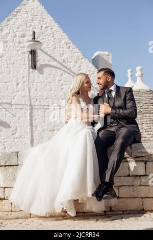 Vertikale romantische verheiratete Paar in der Liebe, blonde Frau und Mann in Anzug nach der Hochzeit, einander suchen, sitzen auf Italien Wand Stockfoto