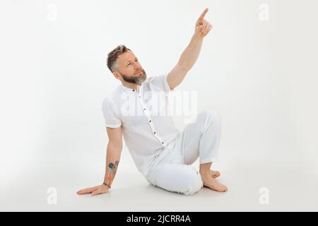 Bärtiger moderner Geschäftsmann sitzt auf dem Boden und präsentiert, zeigt mit dem Finger nach oben, wirbt und wirbt, erklärt den neuen Arbeitsplan Stockfoto