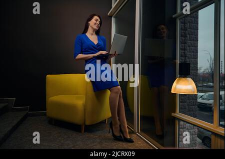 In voller Länge Porträt einer schönen erfolgreichen elegant gekleideten Geschäftsfrau, Unternehmerin, Chefin, die in der Nähe großer Fenster steht und fernab arbeitet Stockfoto