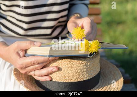 Mittlerer Ausschnitt einer Frau in einem gestreiften Pullover, die in einem Park sitzt und einen Strohhut, ein Buch und einen kleinen Strauß gelber Blumen auf ihren Knien hält. Stockfoto