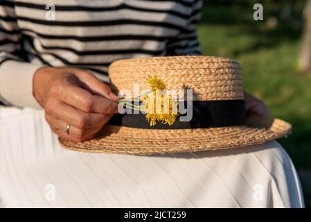 Frau in einem gestreiften Pullover und weißem Rock, die in einem Park sitzt, einen Strohhut und einen kleinen Strauß gelber Blumen auf den Knien hält, in der Mitte geschossen. Stockfoto