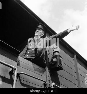 1950s, historisch, ein uniformierte Zugwächter, der in einem hölzernen gerahmten Güterwagen steht und mit einem ausgestrecktem Arm ein Handzeichen gibt, dockt London, England, Großbritannien an. Das Wort Wache ist auf seinen Arbeitshut genäht. An der Außenseite des Wagens hängt eine Bahnleuchte. Stockfoto