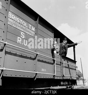 1950s, historisch, ein uniformierter Zugwächter im offenen Heck eines Güterwagens, der mit dem Arm nach außen stand und signalisierte, London Docks, England UK. Schreiben auf der Seite des Güterwagens, Bristol T. M, (Temple Mead) -Paddington. An der Seite des Holzwagens hängt eine Eisenbahnwarnlaterne. Stockfoto