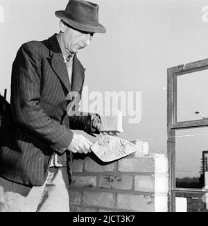 1950s, historischer, älterer Maurer mit Hemd und Krawatte, einer Jacke mit Nadelstreifen und Hut, Kelle in der Hand, Bau einer Wand, England, Großbritannien. Stockfoto