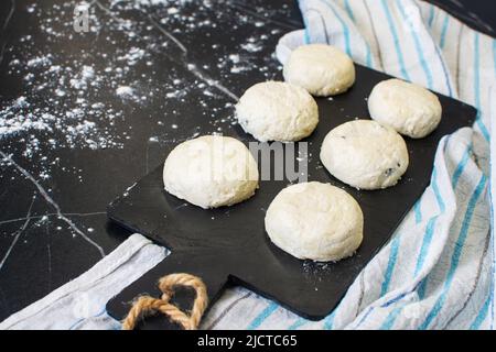 Brett mit ungekochten Hüttenkäse-Pfannkuchen und Zutaten auf schwarzem Hintergrund. Stockfoto