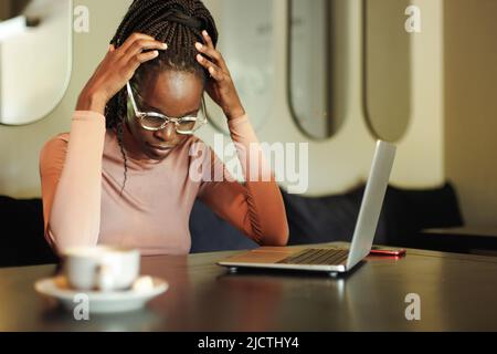 Ängstliche, unselbstbewusste multirassische afroamerikanische Frau in Brille, arbeiten online mit Laptop und Telefon. Kopfschmerzen, Migräne Stockfoto