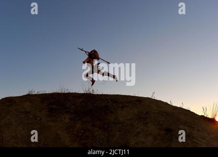 Ein junges Mädchen wird als Indianerin der Ureinwohner Amerikas verkümmelt. Sie wird gesehen, wie sie mit einem Speer in der Hand einen Hügel am Sonnenuntergang hochläuft und anlädt. Stockfoto