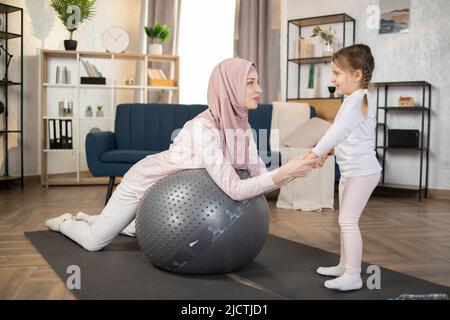 Glückliche, schöne muslimische Mutter in einem Hijab, die mit ihrer kleinen, niedlichen Tochter, die auf einem Fitball liegt und sich gegenseitig die Hand hält, Sport macht. Gesundheit, Schönheit, Sportkonzept. Stockfoto