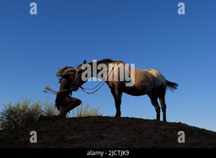 Ein junges Mädchen wird bei Sonnenuntergang als Indianerin der Ureinwohner Amerikas gesehen. Sie hält auf einem kleinen Hügel inne, um ihr Pferd zu schätzen Stockfoto