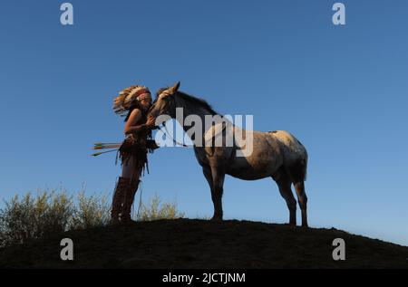 Ein junges Mädchen wird bei Sonnenuntergang als Indianerin der Ureinwohner Amerikas gesehen. Sie hält auf einem kleinen Hügel inne, um ihr Pferd zu schätzen Stockfoto