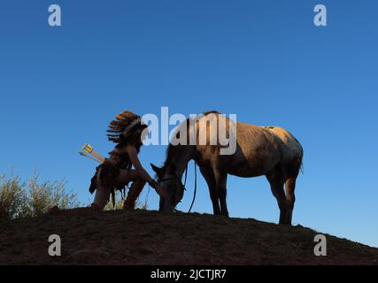 Ein junges Mädchen wird bei Sonnenuntergang als Indianerin der Ureinwohner Amerikas gesehen. Sie hält auf einem kleinen Hügel inne, um ihr Pferd zu schätzen Stockfoto