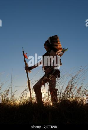 Ein junges Mädchen wird als Indianerin der Ureinwohner gesehen. Stolz posiert sie auf einem Hügel. Das indische Mädchen ist mit der untergehenden Sonne im Hintergrund verkümmelt. Stockfoto