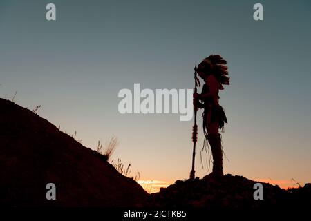Ein junges Mädchen wird als Indianerin der Ureinwohner gesehen. Stolz posiert sie auf einem Hügel. Das indische Mädchen ist mit der untergehenden Sonne im Hintergrund verkümmelt. Stockfoto