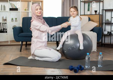 Im mittleren Alter schöne muslimische Sportlerin mit ihrer niedlichen kleinen Tochter tun Yoga-Übung mit Fitness-Ball zu Hause. Junge sportliche arabische Familie lächelt glücklich Training Pilates mit Fitball. Stockfoto