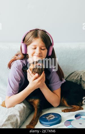 Fröhliches Teenager-Mädchen in lila T-Shirt sitzt auf dem Sofa, hört Musik hat Spaß mit dem Hund spielen Stockfoto