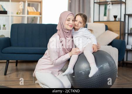 Im mittleren Alter schöne muslimische Sportlerin mit ihrer niedlichen kleinen Tochter tun Yoga-Übung mit Fitness-Ball zu Hause. Junge sportliche arabische Familie lächelt glücklich Training Pilates mit Fitball. Stockfoto