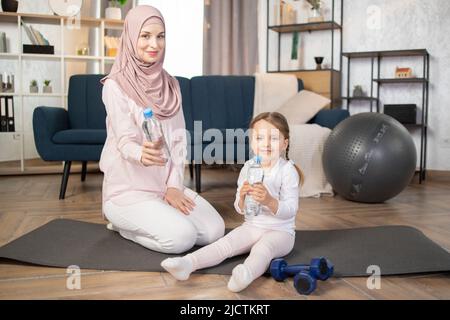 Kleines Mädchen, das mit ihrer jungen muslimischen Mutter auf der Matte sitzt, während sie zu Hause die morgendlichen Übungen macht. Kleines Kind Sport treiben gesunde Praxis von Yoga, Pilates mit Mutter hält Flaschen mit Wasser. Stockfoto