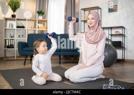 Junge, glückliche arabische Frau, die morgens mit ihrem schönen kleinen Mädchen zu Hause auf einer Matte sitzt und Hanteln in den Händen hält. Stockfoto