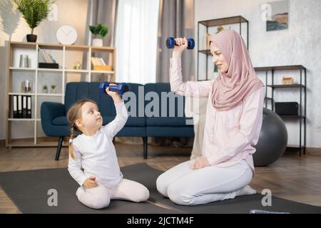 Junge, glückliche arabische Frau, die morgens mit ihrem schönen kleinen Mädchen zu Hause auf einer Matte sitzt und Hanteln in den Händen hält. Stockfoto