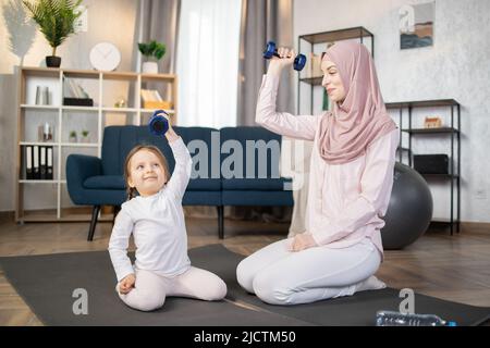 Junge, glückliche arabische Frau, die morgens mit ihrem schönen kleinen Mädchen zu Hause auf einer Matte sitzt und Hanteln in den Händen hält. Stockfoto