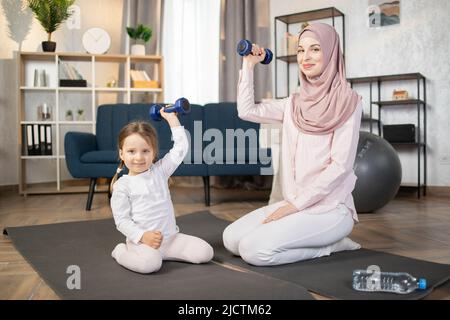 Junge, glückliche arabische Frau, die morgens mit ihrem schönen kleinen Mädchen zu Hause auf einer Matte sitzt und Hanteln in den Händen hält. Stockfoto