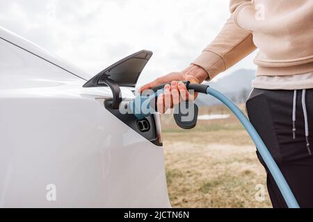 Männliche Hand, die ein Ladegerät in die Steckdose für Elektroautos steckt, im Hintergrund sichtbare Bergnatur Stockfoto