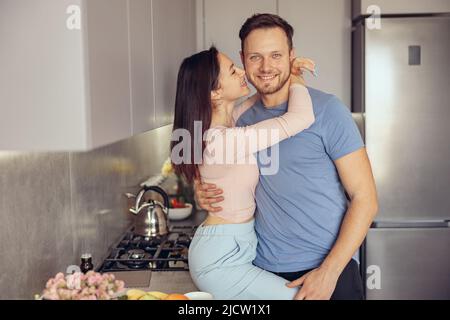 Porträt von glücklichen kaukasischen jungen schönen Mann in der Küche in Umarmungen der geliebten Frau. Stockfoto
