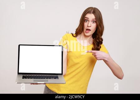 Schockierende Werbung. Porträt eines staunenden Teenagers in einem legeren gelben T-Shirt, das auf einen Laptop mit leerem Bildschirm zeigt und überrascht auf die Kamera schaut. Innenaufnahme des Studios isoliert auf grauem Hintergrund. Stockfoto