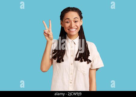 Positive schöne Frau mit schwarzen Dreadlocks zeigt Frieden V-Zeichen, lächelnd zahnig, Gesten Sieg Symbol, mit, gute Laune sorglosen Ausdruck. Innenaufnahme des Studios isoliert auf blauem Hintergrund. Stockfoto